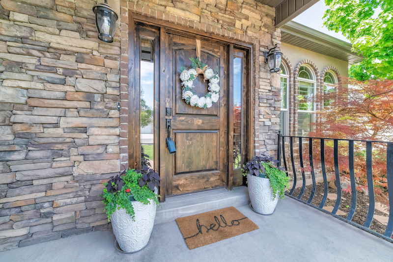 Wooden front door of a house with lockbox and curb appeal. There are two potted plants and doormat at the front of the door with white wreath near the railings against the plants and trees outside.