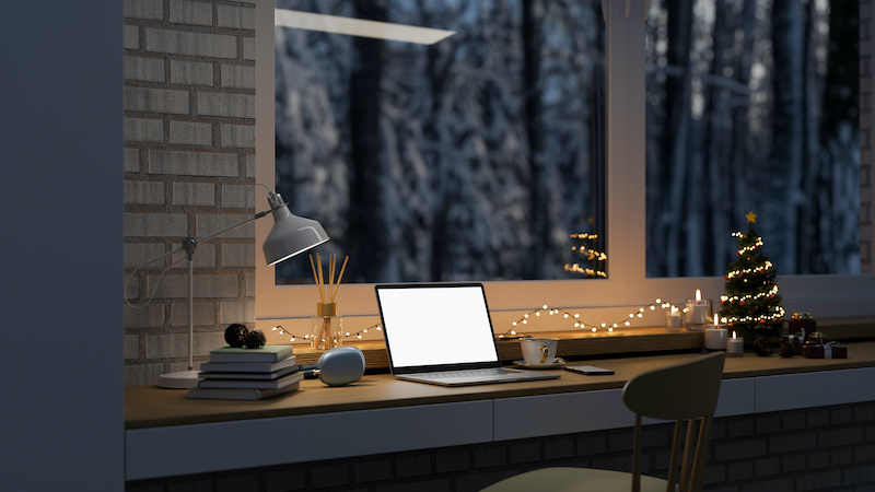 Comfortable home working space with laptop white screen mockup, table lamp Christmas tree, Christmas lights, candles and stuff on table against the window with snow view. 3d render, 3d illustration